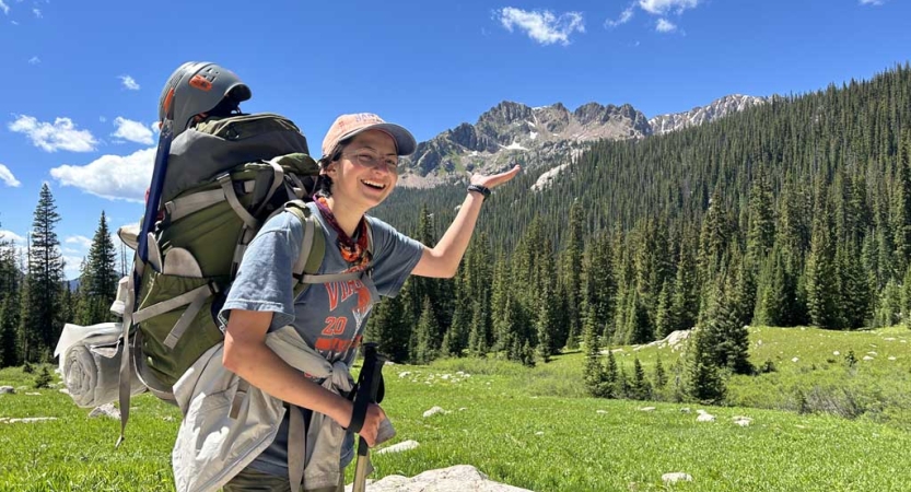 A person wearing a backpack stands in a green alpine meadow and pretends to hold the mountain behind them in the palm of their hand 